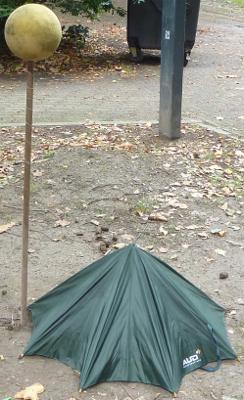 Photo d'un parapluie ouvert sans manche posé sur le sol, à côté duquel est planté un manche de brosse surmonté d'une balle